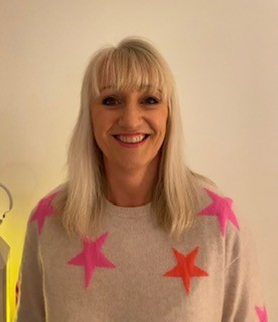 A blond haired woman, wearing a beige jumper with a pink star pattern.