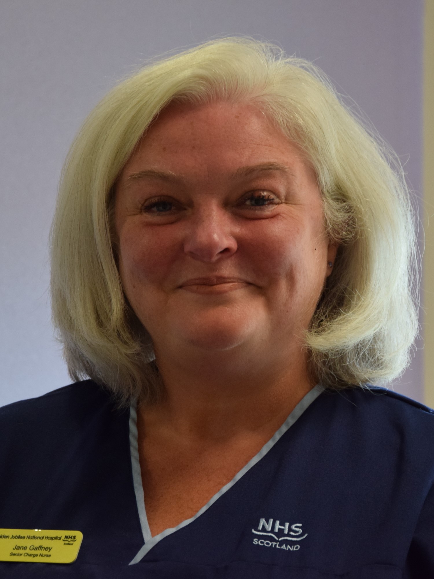 A person with short blond hair, wearing blue NHS scrubs.