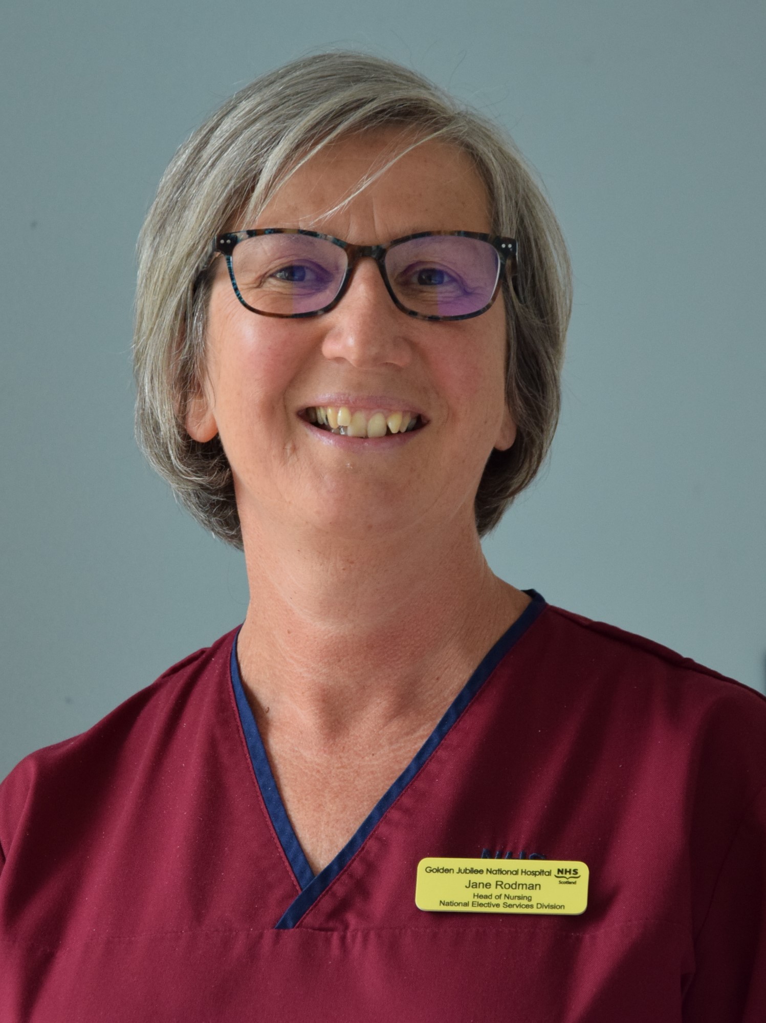 A person with short grey hair, wearing maroon NHS scrubs.