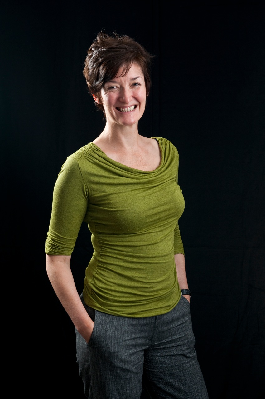 A woman with dark cropped hair, wearing a green top and grey trousers.