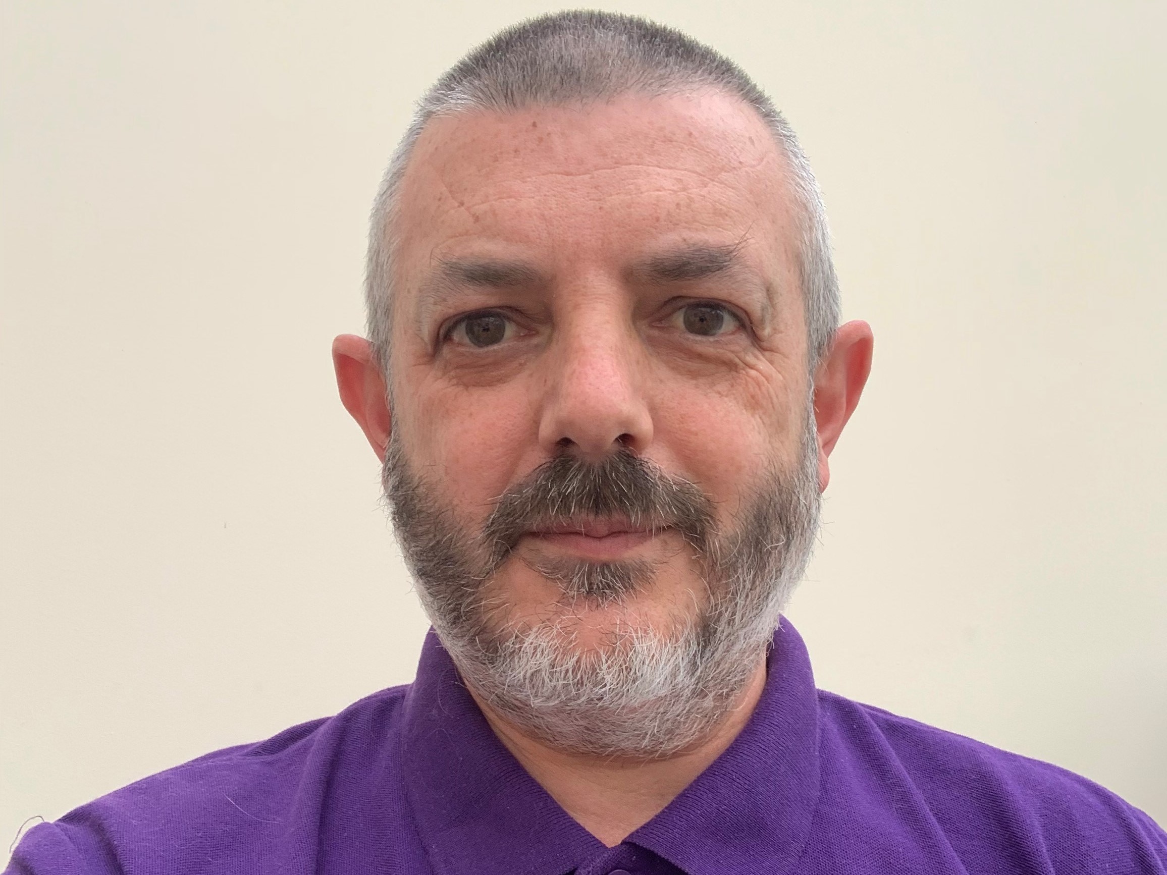 A man with short grey hair and a beard, wearing a purple top.