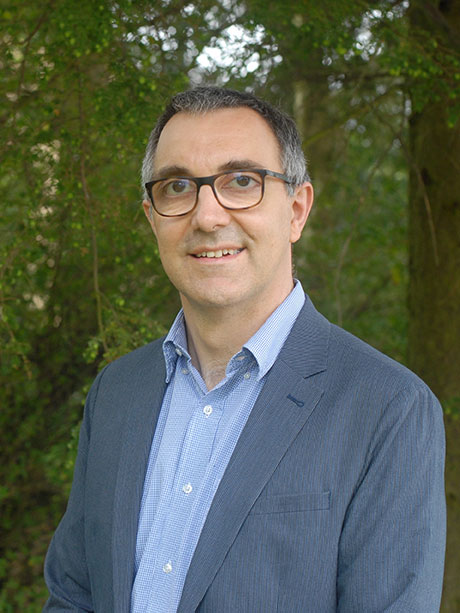 A person with short grey hair, wearing glasses, a grey jacket and blue shirt.