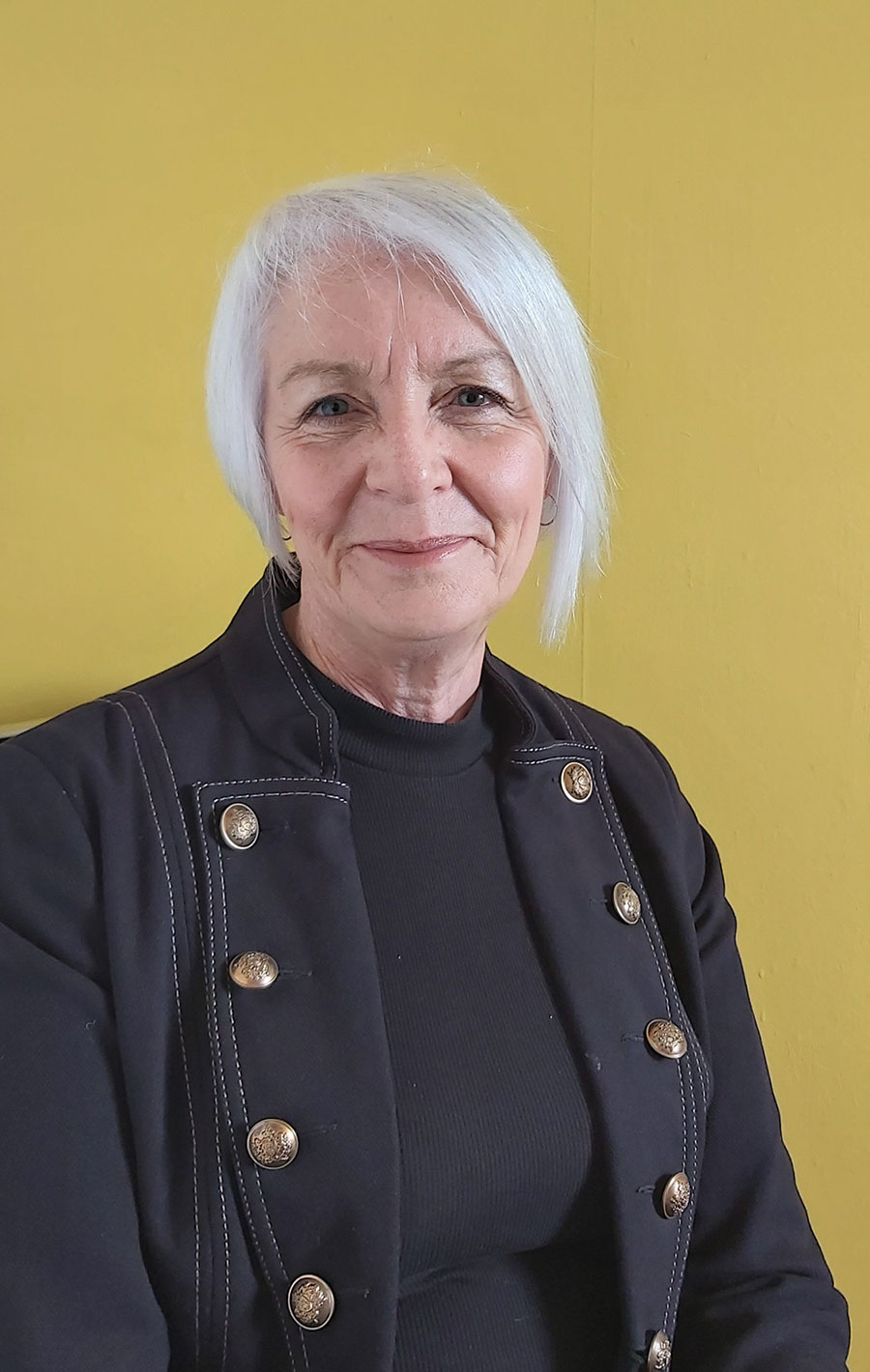 A woman with short grey hair, wearing a blue jacket with buttons.