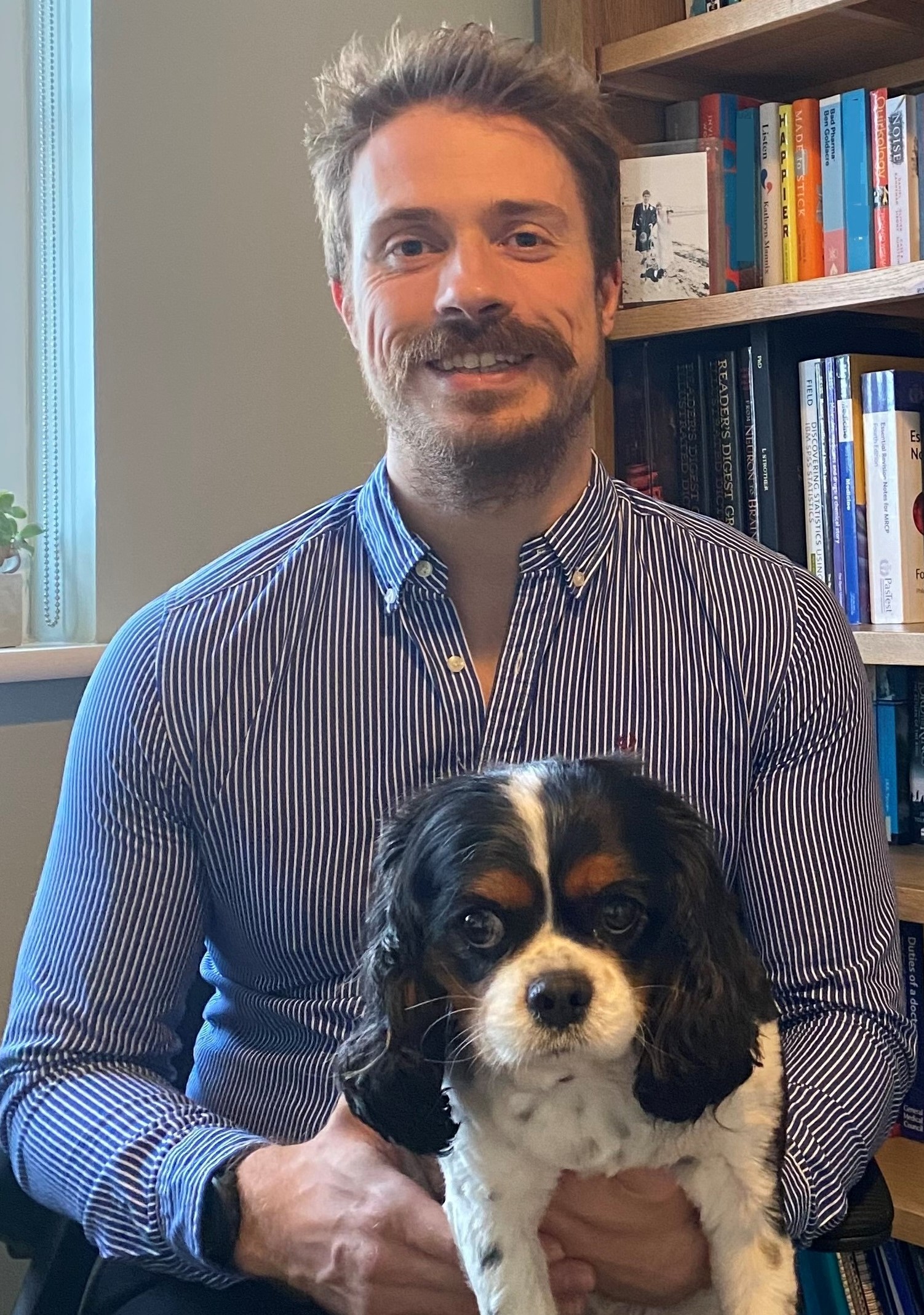 Image of a man with short dark hair, wearing a blue and white striped shirt, holding a spaniel dog