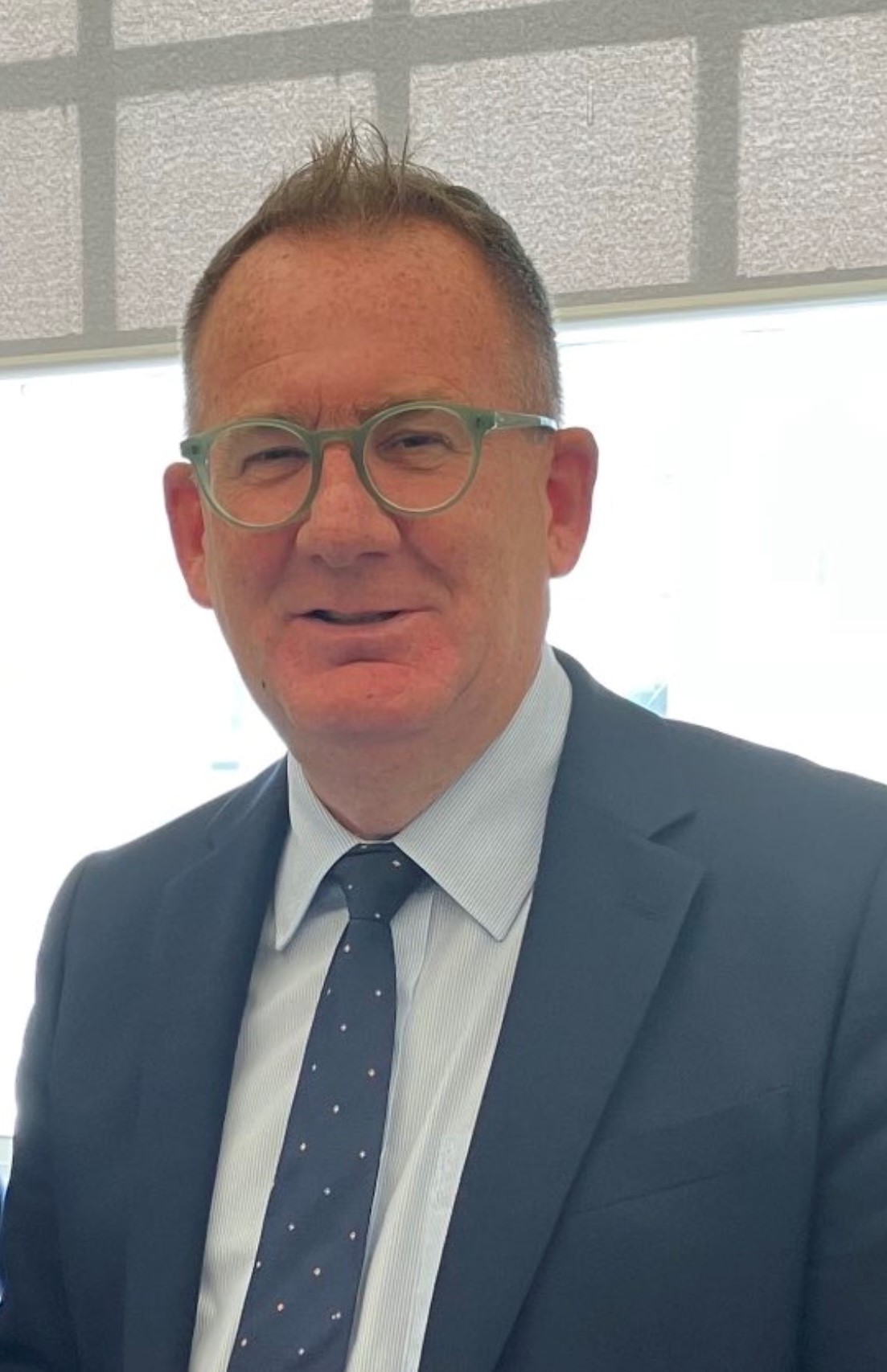 A man smiling, with short grey hair, wearing a light blue shirt, dark blue tie and dark blue blazer