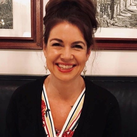 A woman smiling, with dark brown hair, wearing a patterned shirt and a black coat