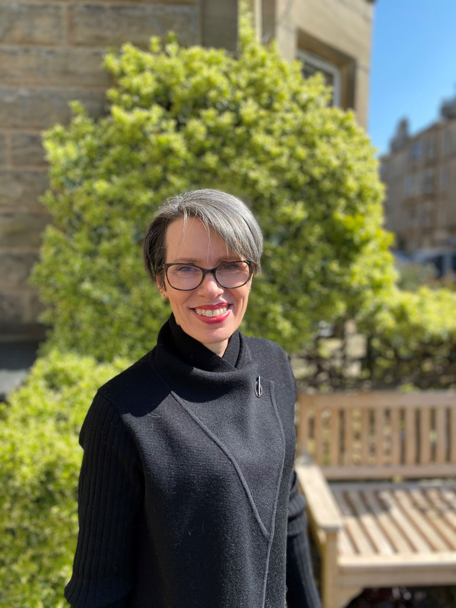 A woman with cropped grey hair, wearing glasses and a black coat.