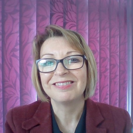 A woman smiling, with short blonde hair and glasses, wearing a red blazer