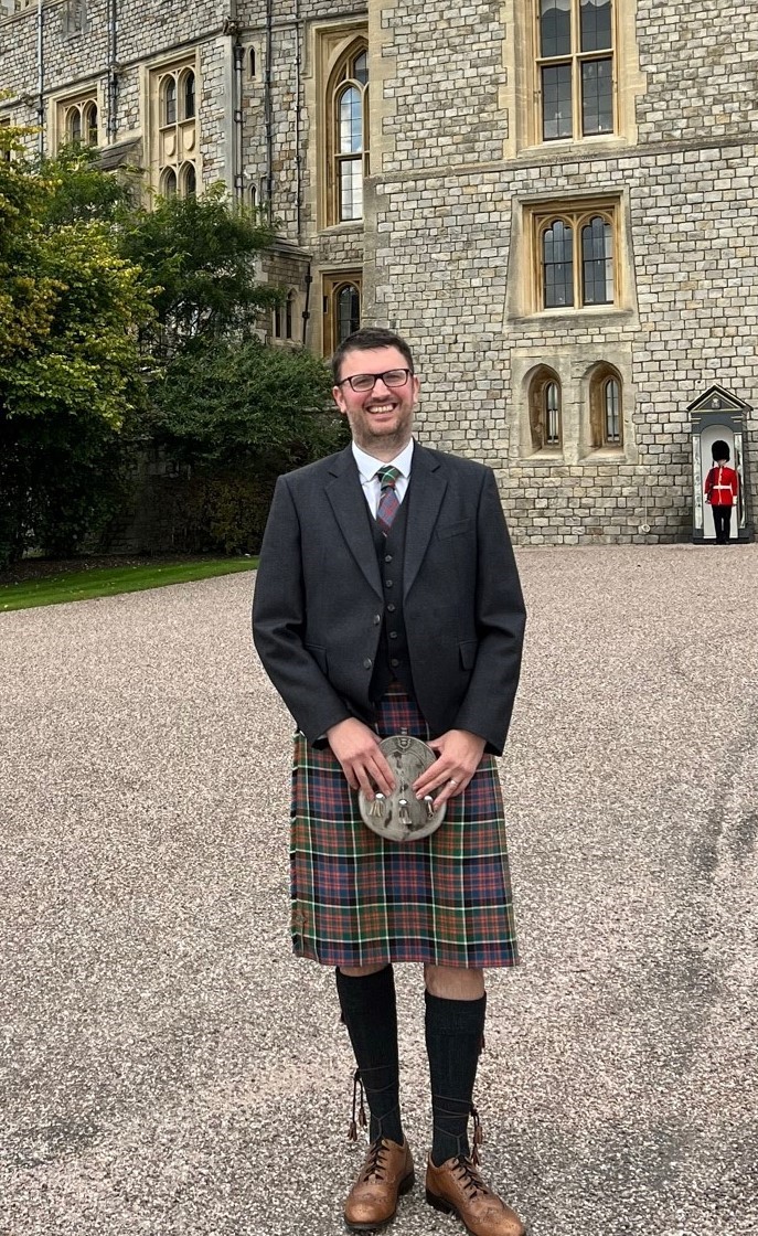 A man smiling, standing in front of a castle, wearing a kilt and blazer