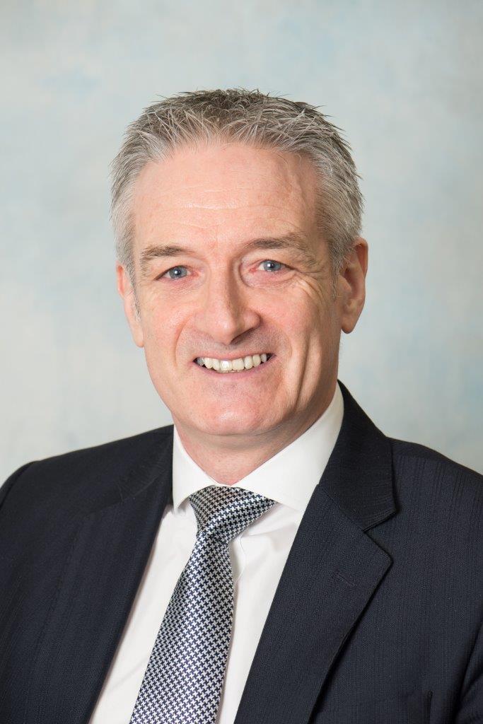 A man smiling, wearing a white shirt, blue tie and a black blazer