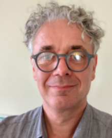 A man with grey curly hair and round glasses.