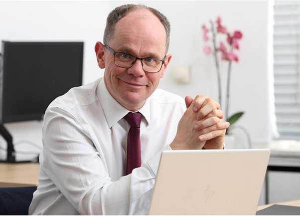 A man smiling, wearing a white shirt with a red tie. There is a pink flower in the background and a laptop in the foreground. 