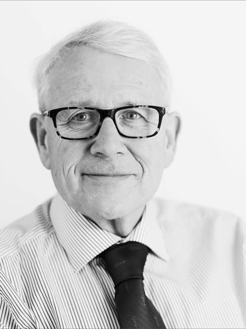 A black and white image of a man with grey hair, black glasses, and wearing a shirt and tie.