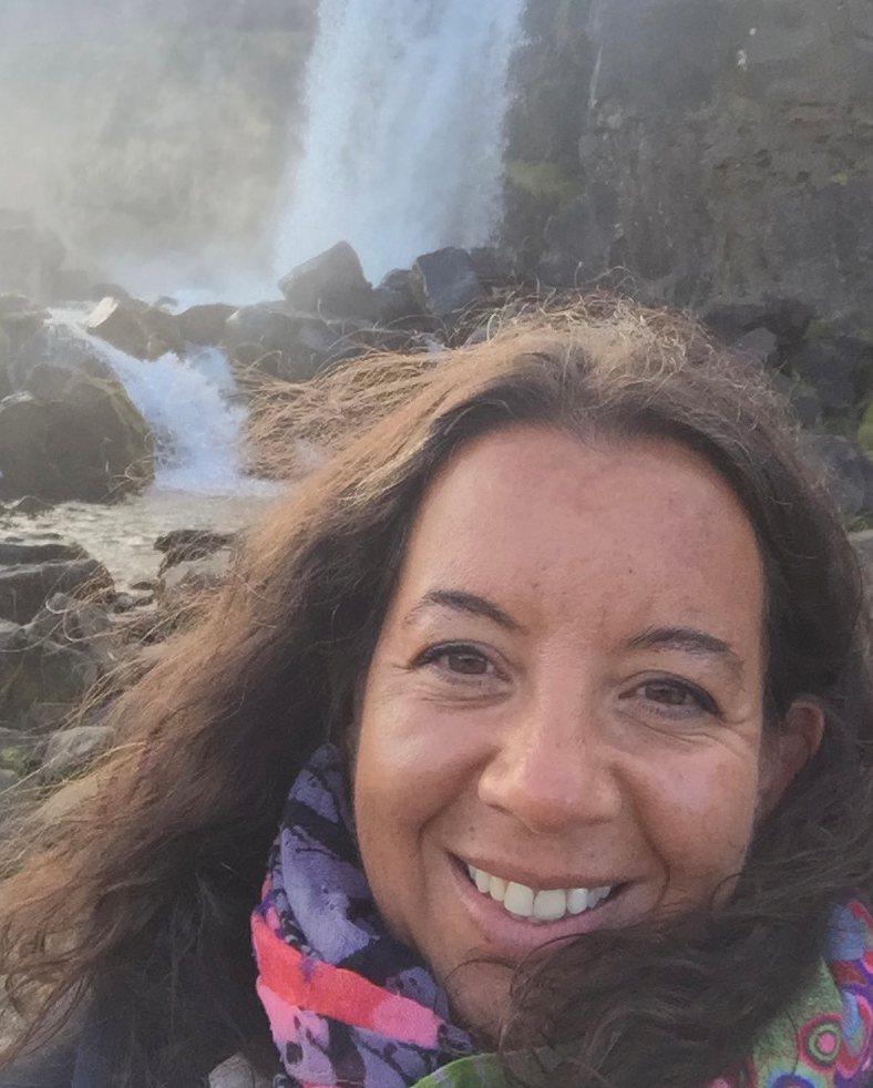 A woman smiling, with long dark brown hair, with a waterfall in the background