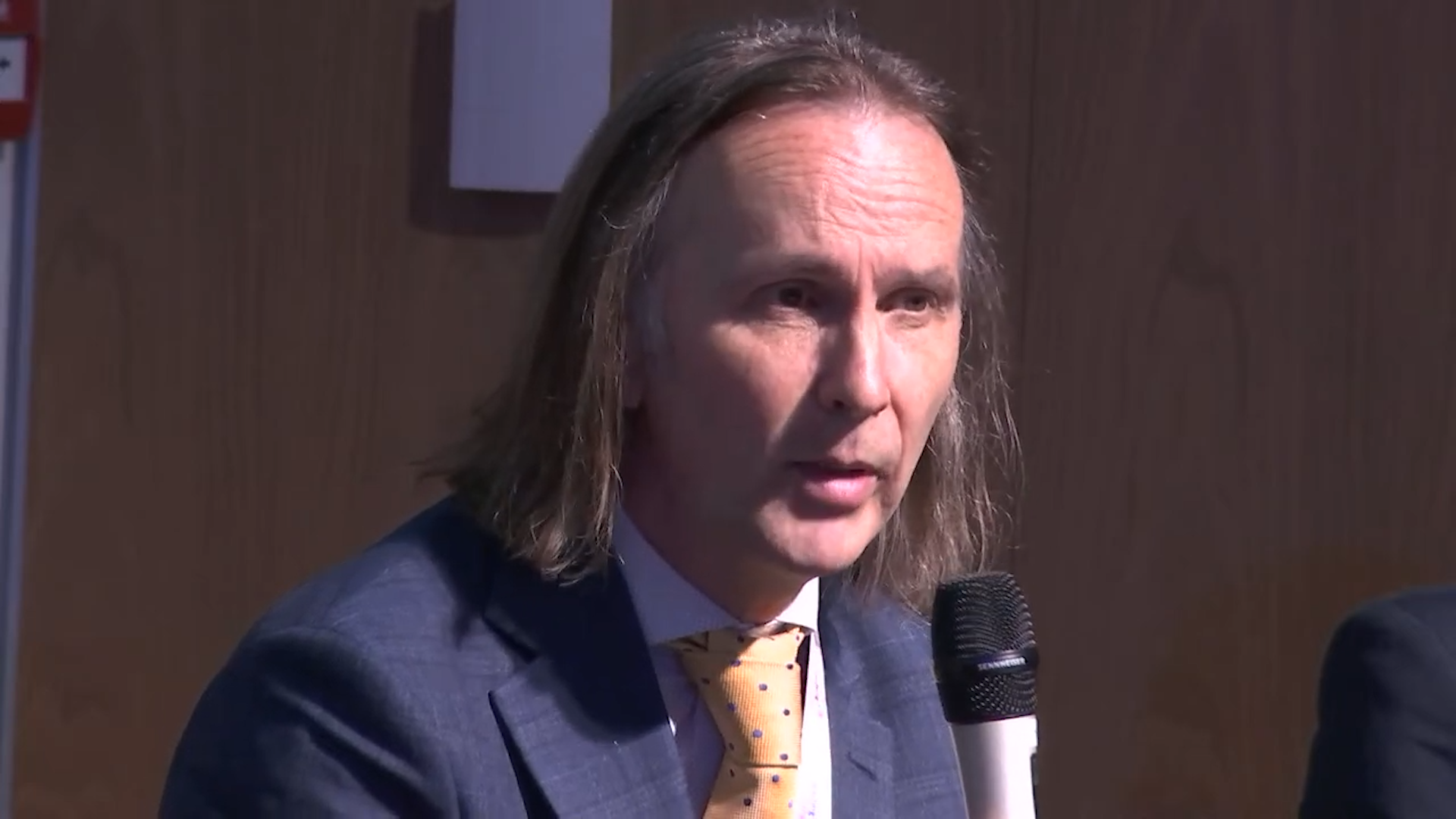 A side-on image of a man wearing a suit and tie, talking at a panel