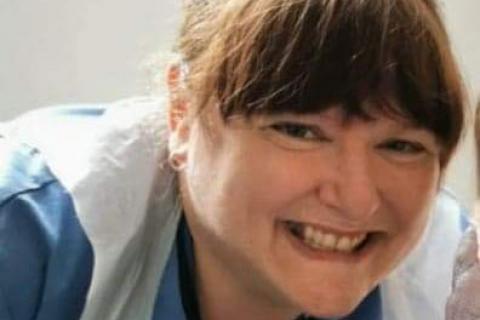 A nurse smiling, wearing a blue uniform and plastic apron.