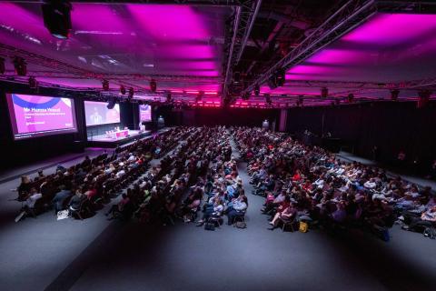 A large event space, with dark pink lighting, a stage and a full audience watching a presentation