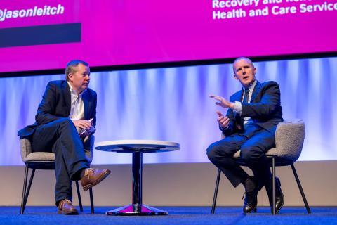 A photo of two men speaking on a stage, sitting down at a small circular coffee table